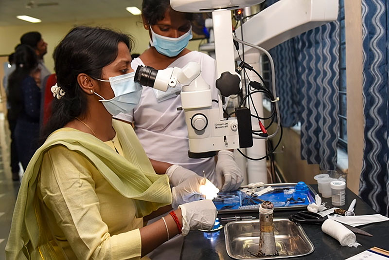 Two female trainees, one is using the surgical microscope, the other stands next to her preparing iinstruments in a tray. Both are wearing blue surgical masks.