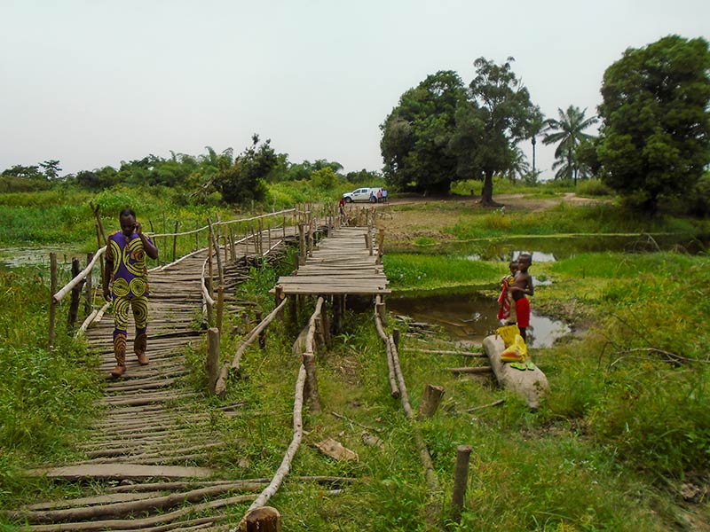 A wooden slatted bridge, one half of the bridge is incomplete. A man uses the walkway and there are two children standing on the river bank close to the broken section.