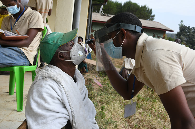A male eye care team member wearing a face visor and mask uses a torch to look at the eye of a male patient who is wearing a face mask