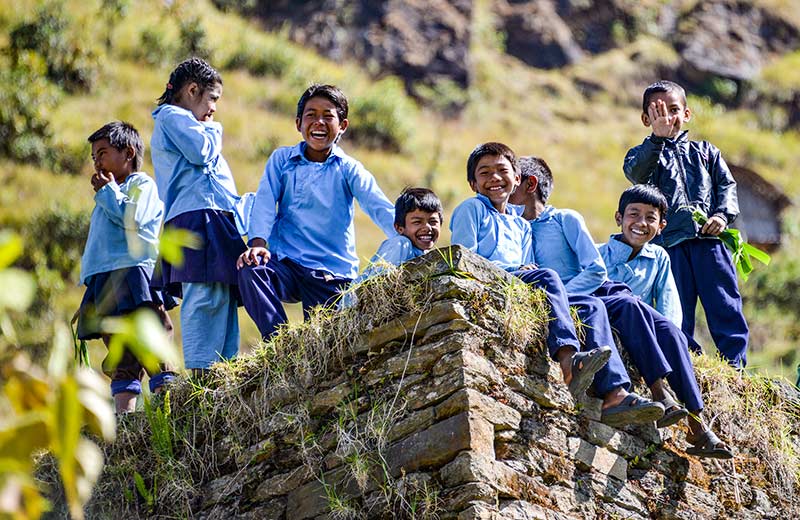Eight children, wearing blue school uniform, sitting or standing on a wall in front of a hillside.