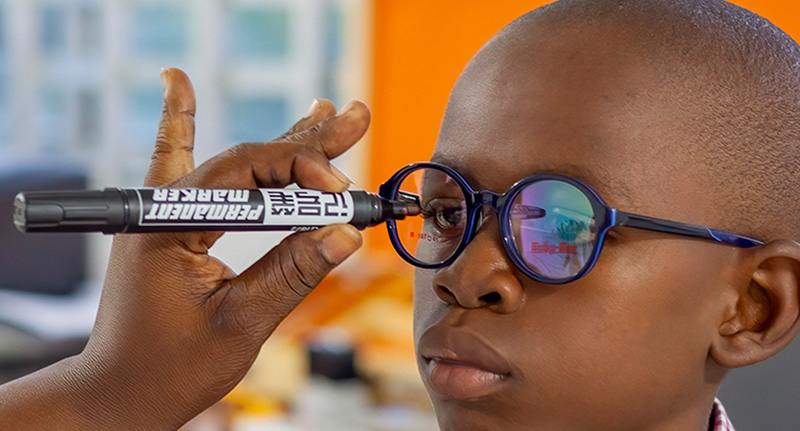 An eye care worker uses a black permanent marker pen to place ad ot on the front of a pair of spectacles to indicate the position of a child's right pupil