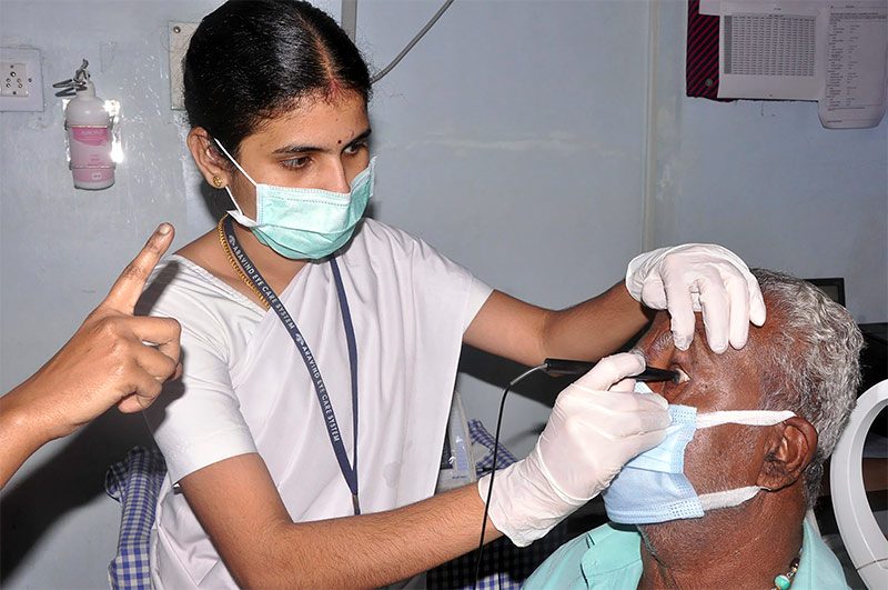 A female biometrist, wearing PPE has her left, gloved hand resting on top of the patient's head to steady it and to hold open his eyelid. She applies the probe to his globe with her right hand.
