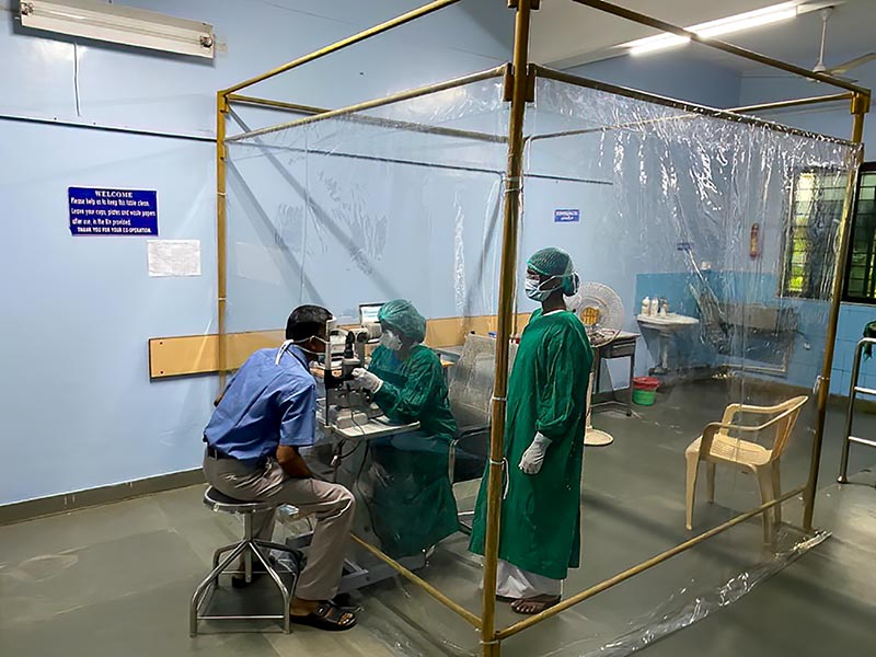 Ophtlamologist and eye nurse standing in a clear plastic screened off area (cube) examining a patient's eye. The patient is outside the protective clear plastic sheeting