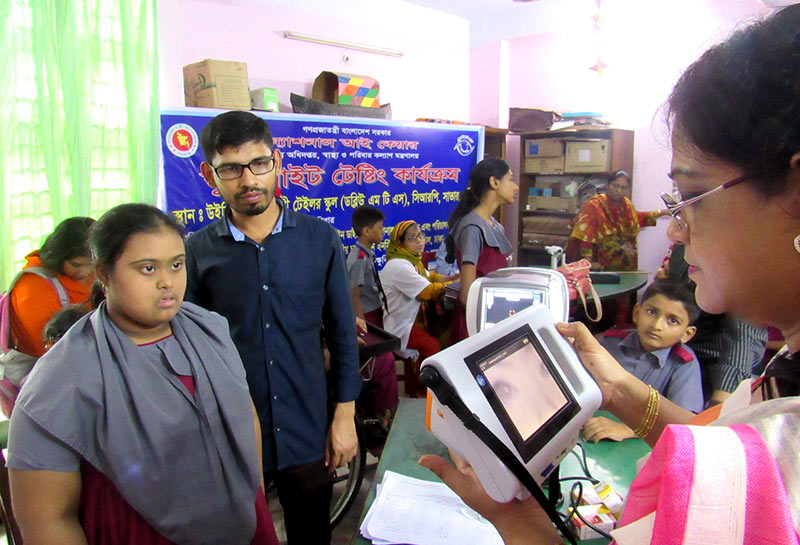 A female eye care worker looks at a digital photograph of the eyes of a young girl with Down's syndrome, whilst standing in front of her. The young girl is accompanied by a male relative.