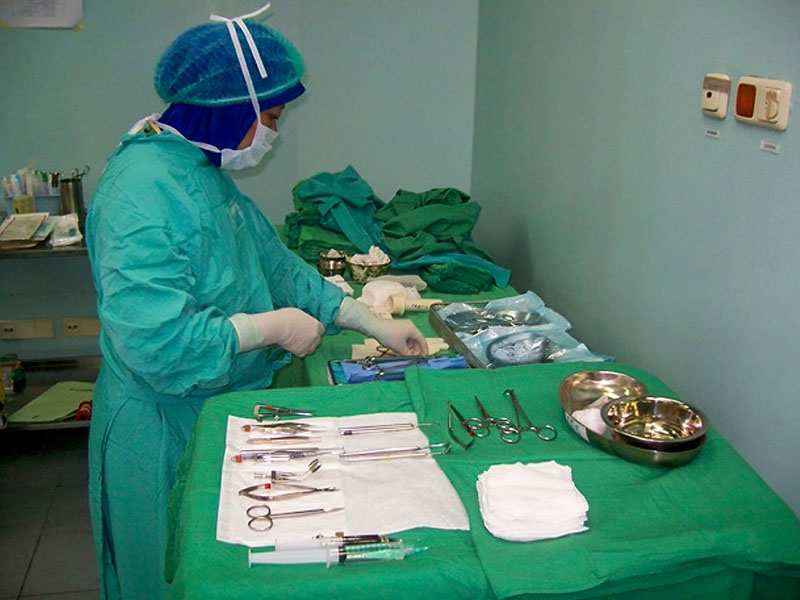 A female staff member dressed in surgical green gown, cap, mask and gloves is picking up items from a tabel covered with a green drape. The table/trolleys have lots of surgical instruments laid out on them.