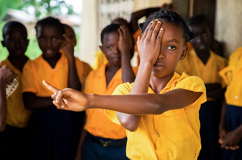 School-age children dressed in bright yellow shirts. A girl at the front of the group is covering her right eye with her right hand and pointing right with her left hand.