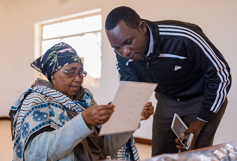 A male eye care worker assists a female patient. The female patient is wearing spectacles and she is reading from a white sheet or card/paper.