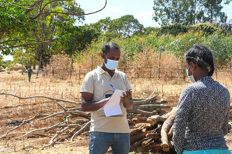 A male health worker wearing a mask writes down notes with a a biro onto a pad of paper. He is standing in front of a female health worker who is also wearing a mask.