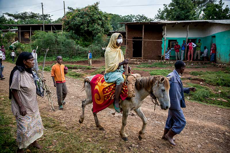 A gentleman leads a donkey though a village carrying a lady wearing eye patches