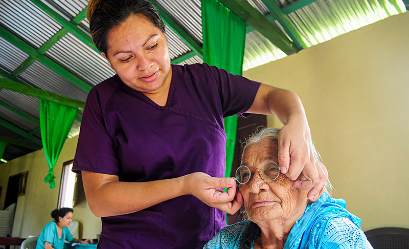 A female health care worker wearing purple scrubs places spectacles on an elderly woman's face.