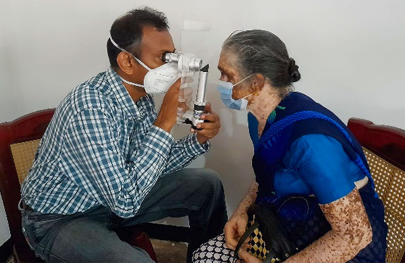 A male ophthalmologist, who is wearing a face mask, uses a portable slit lamp with a protective screen, to examine the eyes of a female patient (she is also wearing a face mask).