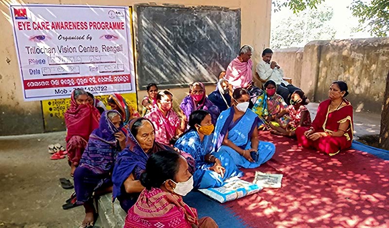 A group of female volunterrs are sitting on a rug, outside a building in a walled garden area. There is a poster about the eye care awareness programme on the wall behind them.