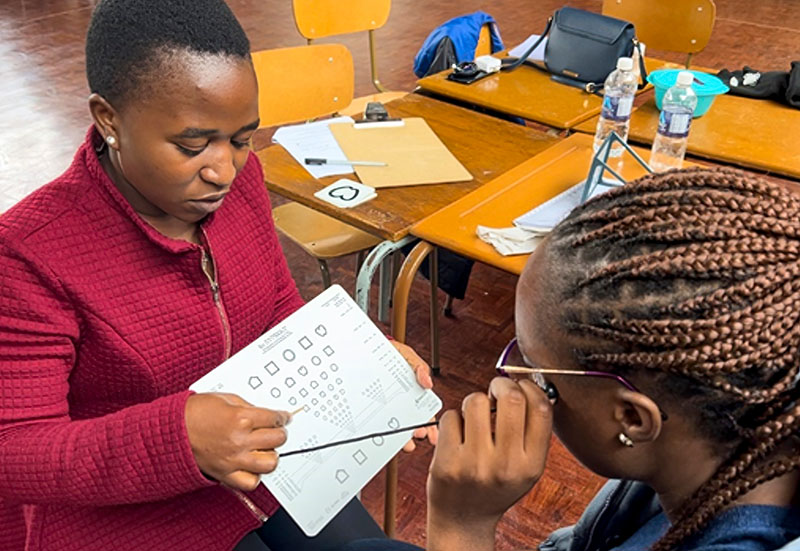 A female eye health worker is pointing to a chart she is holding in front of a female patient who is wearing glasses. Both people in the image are seated.