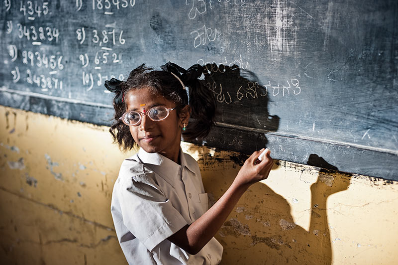 Une écolière portant des lunettes écrit sur un tableau noir avec de la craie, dans une salle de classe.