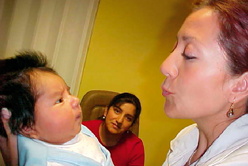 Une agente de santé oculaire tient un nourrisson qui regarde son visage, pendant que la mère de l’enfant est assise et les regarde.