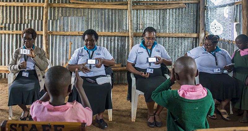 Four school teachers sit opposite school children. Each of the teachers is holding a smartphone displaying the Peek acuity app.