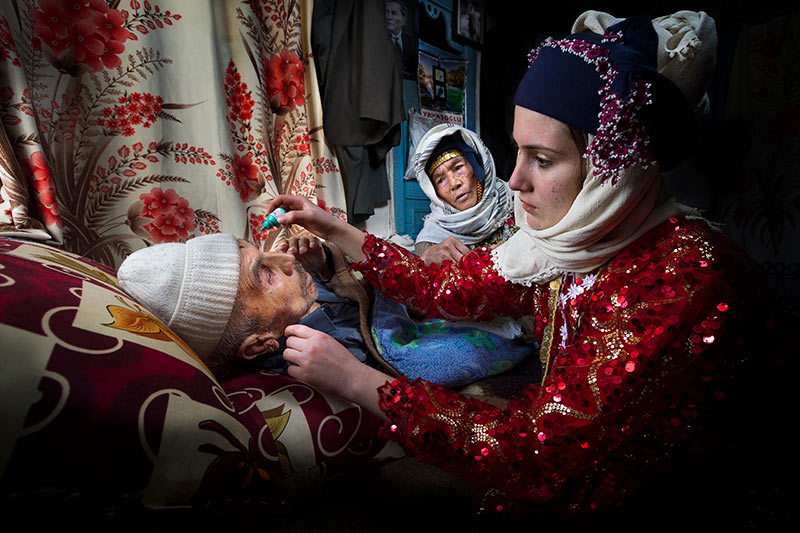 A young woman instills eye drops into her father's eyes while he is lying down. Her mother watches from the side of the bed.