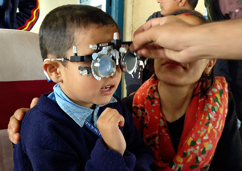 the arm of an eye health worker reaches out to place a pair of trial lenses on a young boy whose mother is kneeling next to him with her arm around him