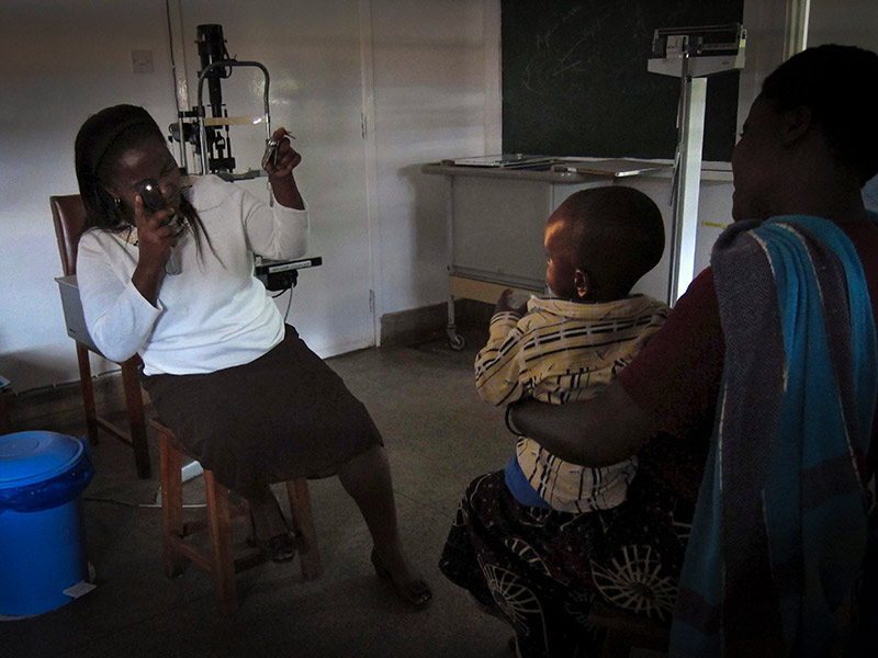 Dans une pièce sombre, un agent de santé tient un instrument pour examiner le reflet pupillaire d’un enfant, qui est assis à une certaine distance, sur les genoux d’un parent.