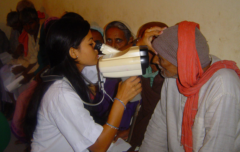 a female ophthalmic assistant holding a handheld refraction device up to the eyes of an elderly gentleman sitting down in the hallway next to other patients who are waiting