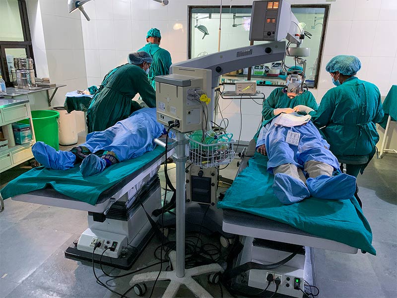 Two patients on operating beds in theatre. The ophthalmic surgeon is operating on the patient on the left assisted by an theatre nurse and two clinical staff prepare the other patient.
