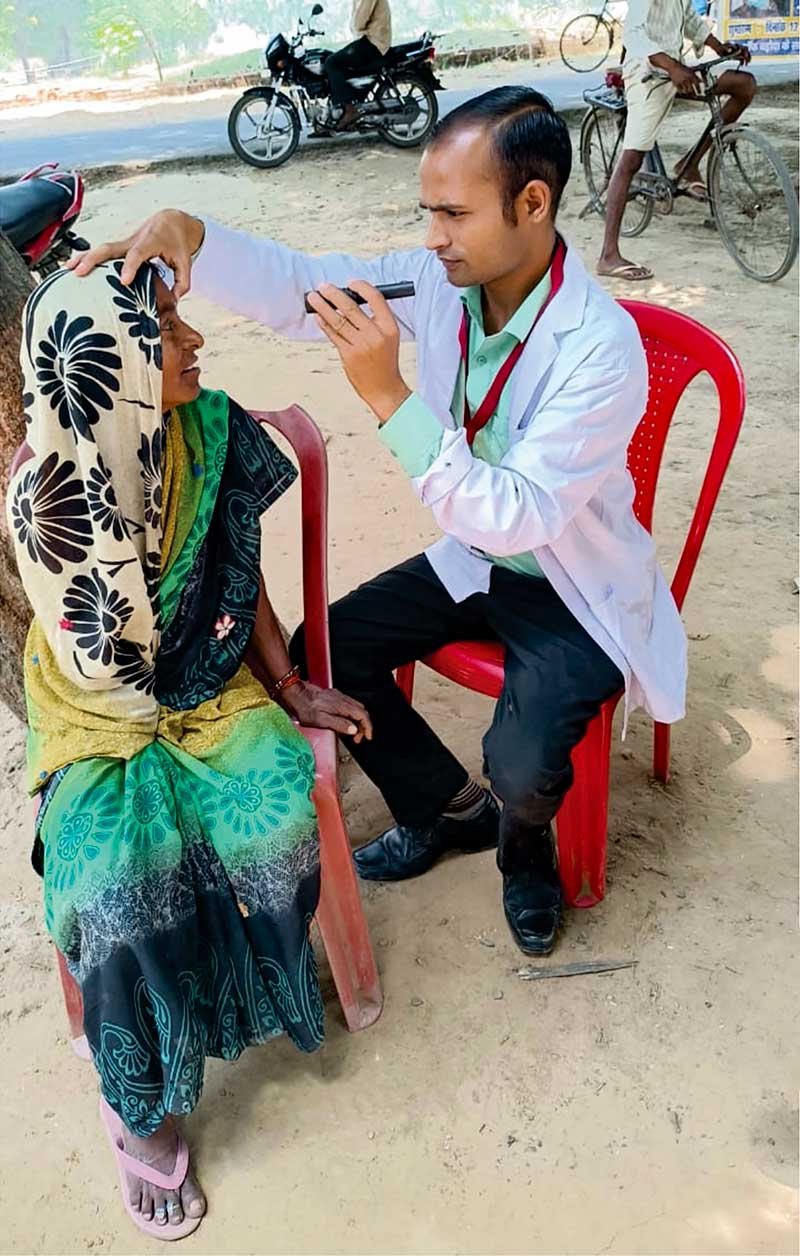 A male health worker examines the eyes of a female patient with a torch. Both are sittign down on red chairs outside.
