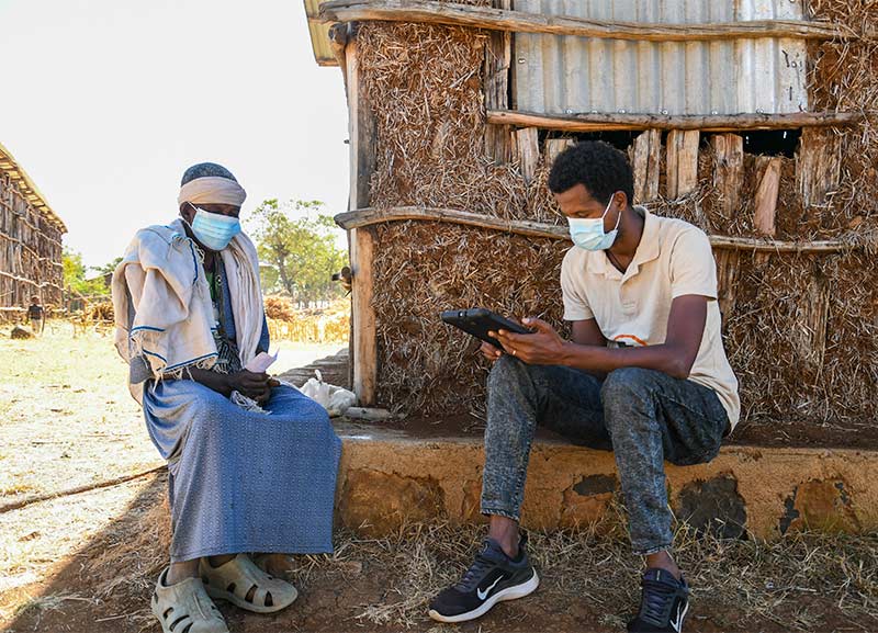 A male health worker sits with a male patient outside a house, entering data into an i pad.