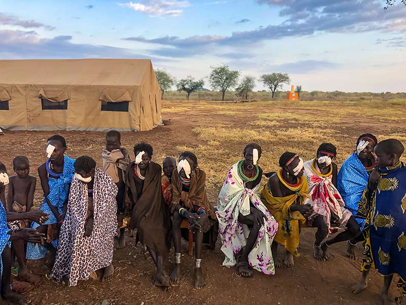 Adult patients wearing eye bandages post-surgery and their children sitting in front of a large brown tent.
