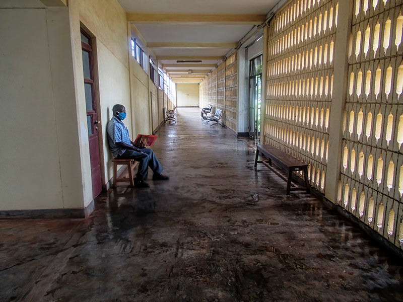 Male patient, wearing a face mask, sitting alone on a bench in a hospital corridor.