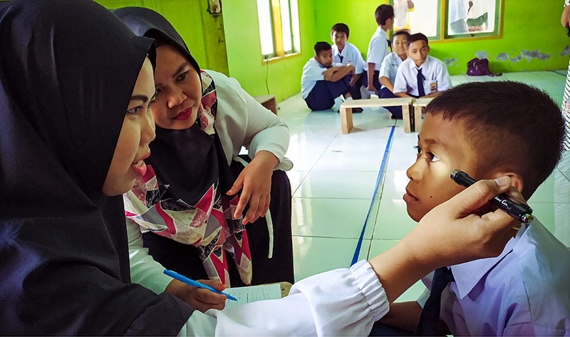 Two female eye care workers examining a boy's eyes in a classroom using a torch
