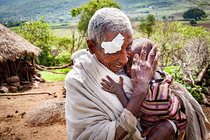 An elderly man standing outside his thatched house holding a child in his arms. The man is wearing an eye patch bandage and smiling.