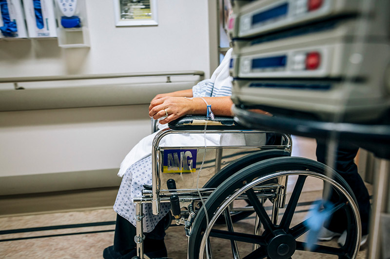 A patient sits in a wheelchair partially hidden by hospital monitoring equipment