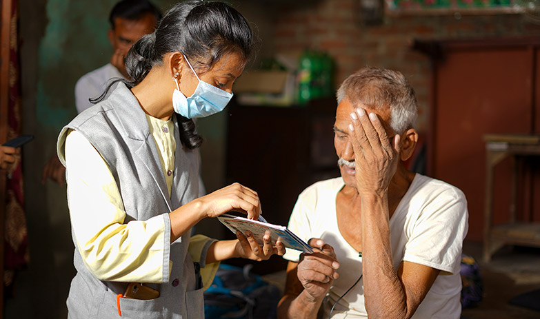 a female health worker points to a gook she is holding with a pen while standing close to an elderly gentleman who is covering his left eye with his left hand
