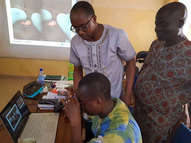 A male student uses 3-D glasses to view a computer laptop screen. A male tutor stands next to him explaining the activity and a female student is also standing with them watching.